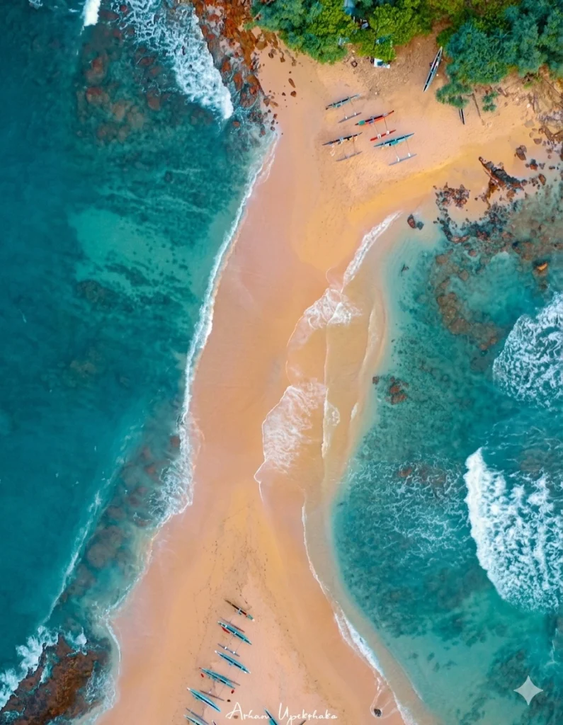 Aerial view of a narrow sandbar flanked by turquoise ocean waves and colorful outrigger boats parked on the golden shore.