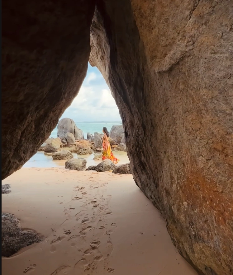 A traveler in a vibrant orange dress walking on a sandy beach, viewed through the opening of a rocky sea cave.